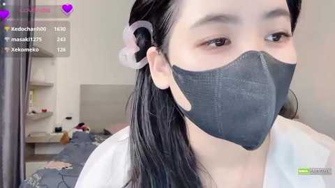 Media: Video of an Asian woman with long black hair tied back, wearing a grey face mask, white shirt, and scrunchie, in a modern bedroom with a bed, white walls, and purple chat window.