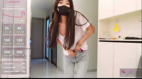 A video of an Asian woman with long brown hair, wearing a black mask and white top, adjusting her pants, in a modern kitchen with white cabinets.