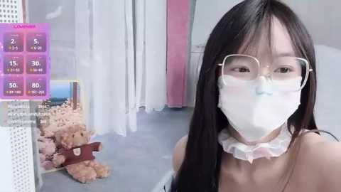 Video of an Asian woman with long black hair, wearing glasses and a surgical mask, standing in a store with teddy bears and a virtual reality display.
