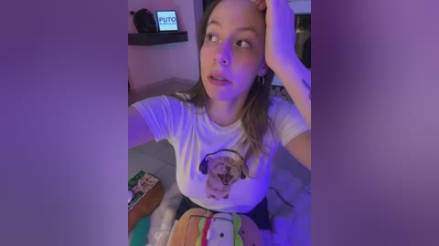 Video of a young woman with light skin, light brown hair, wearing a white t-shirt with a dog graphic, sitting on a bed, holding a phone, and looking contemplative under purple lighting.