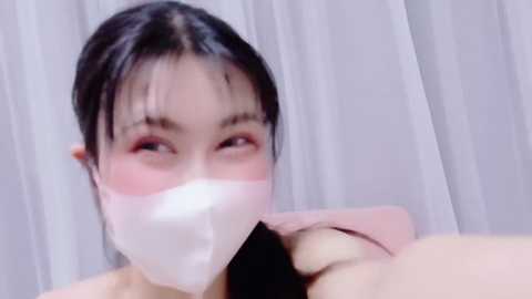 Media: Video of a young Asian woman with fair skin, dark hair, and light pink lipstick, wearing a white face mask, performing oral sex on a man. The background features white curtains.