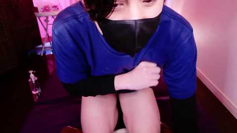 Video of a person wearing a blue jacket, black mask, and black gloves, with a bottle nearby, kneeling on a dark floor, in a room with pink lighting.