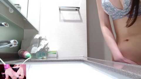 A video of a woman in a bra and pink robe standing in a modern bathroom with white walls and a sink, partially reflecting her face in the mirror.