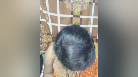 Video of a woman with black, voluminous hair sitting on a chair. She wears an orange dress with white and brown patterns. The background features a brown curtain with intricate designs.