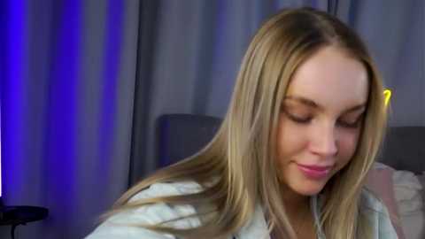 Video of a smiling young woman with long blonde hair, wearing a white floral-patterned shirt, sitting in a dimly lit room with grey curtains and a blue accent light.