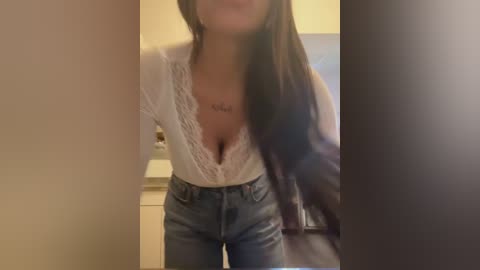 Video of a light-skinned woman with long dark hair, wearing a white lace-trimmed blouse revealing cleavage, and high-waisted blue jeans. She stands in a dimly lit room with blurred background.