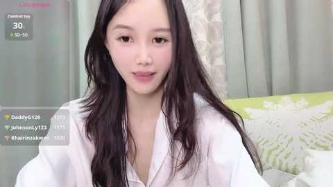 Video of an East Asian woman with long black hair, fair skin, and a slim build, wearing a white robe, sitting on a bed with a green floral-patterned pillow.