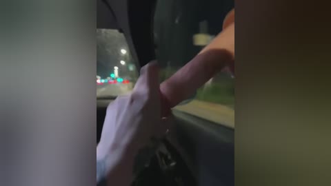 A dimly-lit video captures a hand gripping a penis inside a car, with blurred streetlights visible through the window. The setting appears to be nighttime, and the image is slightly out of focus.