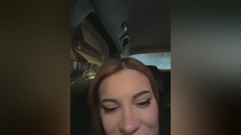 Video of a young woman with fair skin, light brown hair, and light makeup, seen from the neck up, in the dimly lit interior of a car, with a soft focus and slightly blurred background.