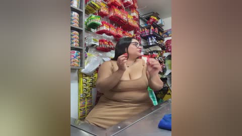 Video of a plus-sized woman in a beige dress, holding a phone, in a crowded convenience store aisle packed with snacks and candy.