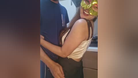 Media: Video of a smiling Asian woman in a yellow mask, wearing a white top and black skirt, hugging a man in a navy shirt, in a kitchen with tiled walls.