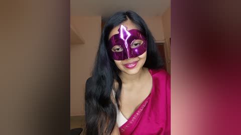 Video of a smiling woman with long black hair, wearing a vibrant pink saree and a shiny purple mask with a white design, standing indoors against a beige wall.