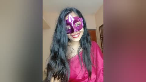 Video of a smiling woman with long black hair, wearing a vibrant pink satin dress and a purple Venetian mask. Background shows a plain, beige wall and part of a wooden door.
