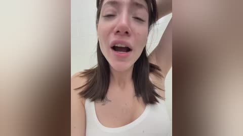 Video of a young, fair-skinned woman with straight brown hair, wearing a white tank top, eyes closed, mouth open, possibly in pain or ecstasy, blurred background.