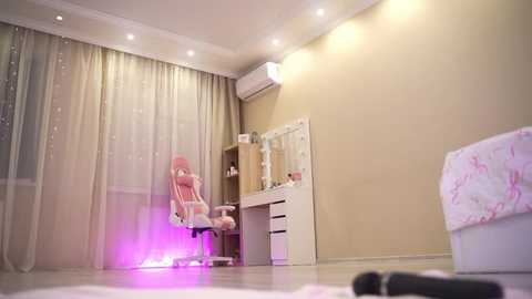 Video of a pink gaming chair in a dimly lit, beige-walled room with sheer curtains, a desk, and a bed with pink bedding.