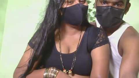 Video of a dark-skinned woman with long black hair and a black face mask, wearing a black top, gold necklace, and bracelets. A dark-skinned man in a white tank top stands behind her, also masked. Both are indoors with a pale green wall background.
