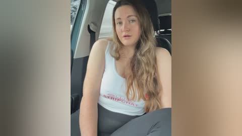 Video of a young woman with long, wavy brown hair, wearing a white Hooters tank top and gray leggings, sitting in a car with a blurred background.