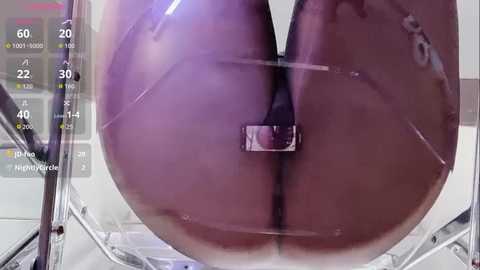 A video of a person sitting on a clear acrylic chair, revealing their bare buttocks and genital area through the chair's transparency. The background shows a digital interface with health and fitness data.