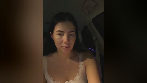 Video of a young Asian woman with straight black hair, wearing a white lace bra, seated in a dimly lit car. She has a serene expression.