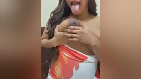 Video of a woman with long dark hair, light brown skin, and a tongue sticking out, covering her bare breast with her hands. She wears a white dress with red floral print.