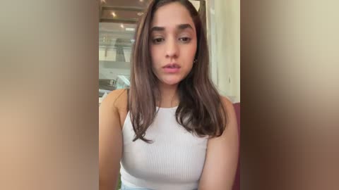 Video of a young Latina woman with long brown hair, wearing a white ribbed tank top, sitting indoors. Background features a glass window and modern decor.