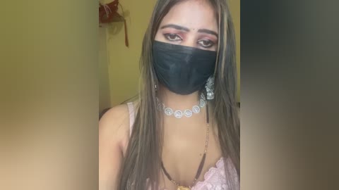 Video of a young woman with light brown skin and long, straight, brown hair. She wears a black face mask, pink lace bra, and a silver necklace with circular pendants. The background is a yellow wall with a small, red shelf.