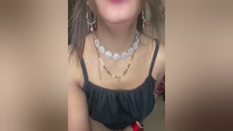 A video of a woman with fair skin and long brown hair, wearing a black camisole and intricate silver necklace with red beads, set against a blurred background.