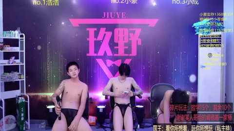 Video of two shirtless Asian men in black briefs, seated on a stage with a neon pink backdrop. One man is facing forward, while the other is facing away.