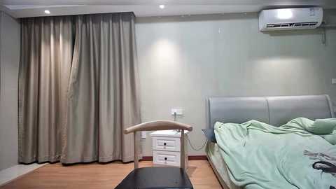 A video of a minimalist, modern bedroom with light green walls, a gray headboard, beige curtains, white bedside table, and an air conditioner.