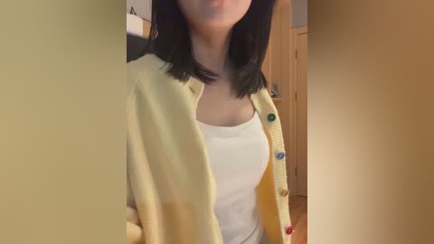 Video of a woman with shoulder-length black hair, wearing a white tank top and a yellow cardigan with colorful buttons, standing in a hallway with beige walls and a wooden door.