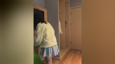 Video of a young woman with black hair wearing a light sweater and blue skirt bending over to pick something up in a small, dimly lit room with wooden floor and light blue walls.
