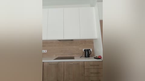 Video of a minimalist kitchen with white cabinets, light wood countertops, beige tile backsplash, a black kettle, and a red apple on the counter.