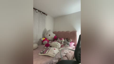 Video of a small, dimly lit room with white walls and a pink quilted bedspread. Various plush toys, including Hello Kitty and other characters, are scattered on the bed, creating a cozy, child-like atmosphere.