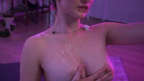 Video of a topless woman with light skin and medium-sized breasts, wearing a heart-shaped pendant necklace. She has a shiny, wet appearance, with a trail of liquid running down her chest. Background features a purple-lit room with plants.