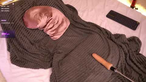 A video shows a person lying on a bed with a black knit blanket, wearing a pink eye mask and a wooden-handled screwdriver placed next to them. The room is dimly lit with a keyboard and computer monitor visible in the background.