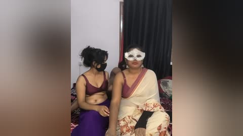 A video of two women in traditional Indian attire, one in a purple top and purple pants, the other in a white sari with red patterns, sitting on a bed with black curtains in the background.