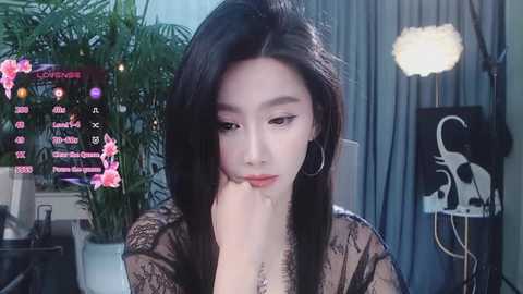 Media: Video of an East Asian woman with long black hair, wearing a black lace top, sitting indoors with plants, a monitor, and a spotlight in the background.