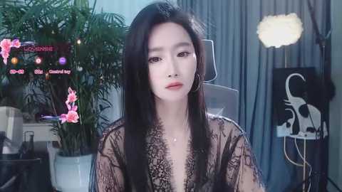 Media: Video of a young Asian woman with long black hair, fair skin, and full lips, wearing a sheer black lace robe, seated in a modern room with a green plant, a light bulb, and a chair.