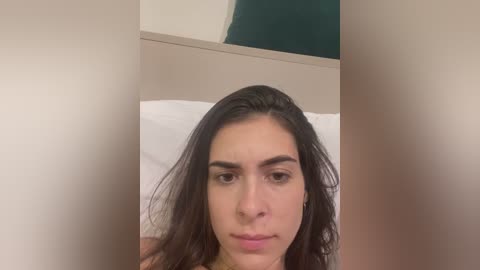 Video of a young woman with long, dark hair, fair skin, and dark eyebrows, wearing a light-colored top. She appears contemplative with a neutral expression, sitting on a bed with white pillows and a beige headboard in a minimalist room.