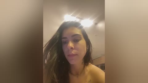 Video of a young woman with medium-length, wavy brown hair, light skin, and full lips, wearing a strapless top. She stands indoors under a ceiling light. The background is a beige wall.