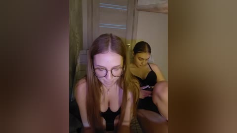 Media: Video of two women in a dimly-lit room. A blonde with glasses, wearing a black bra, leans forward, looking down. A brunette with a ponytail sits beside her, also in a black bra, holding a phone.