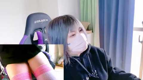 A video of an Asian woman with light skin, wearing a black mask, purple and pink socks, and a black jacket, sitting in a gaming chair with a blue and green curtain in the background.