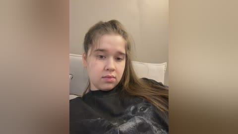 Video of a young Caucasian girl with long brown hair, wearing a black cape, sitting on a white couch, looking pensive, in a beige-walled room.