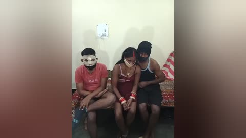 Video of three bound individuals, two men and one woman, wearing masks, seated on a couch in a dimly lit room with a white wall and a red blanket.