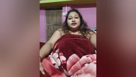 Video of a plus-size woman with light brown skin, wearing a red saree with a floral pattern, sitting on a bed with a pink blanket. Background shows a wooden door and pink wall.