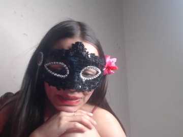 Video of a woman with long black hair, wearing a black lace masquerade mask with pink flowers, holding her hands together, smiling.