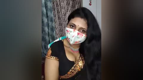 Video of a South Asian woman with long black hair, wearing a floral face mask, black saree, and silver jewelry, standing in a room with patterned curtains and a white door.