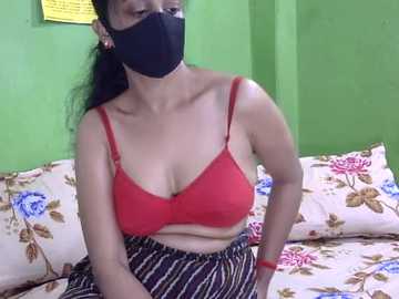 Video of a South Asian woman with medium skin tone wearing a black face mask, red bra, and striped skirt, sitting on a floral bedspread in a green-walled room.