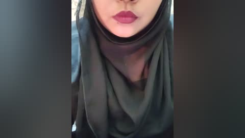 A video of a woman's face, focusing on her lips painted in deep red lipstick, partially covered by a black, sheer hijab. The background is blurred, with soft lighting enhancing the soft texture of the hijab.