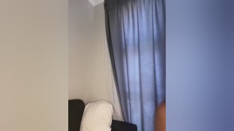 Video of a small, dimly-lit bedroom corner. A white pillow rests on a dark brown headboard against a light beige wall. A gray curtain partially covers a window, casting a blueish light into the room.